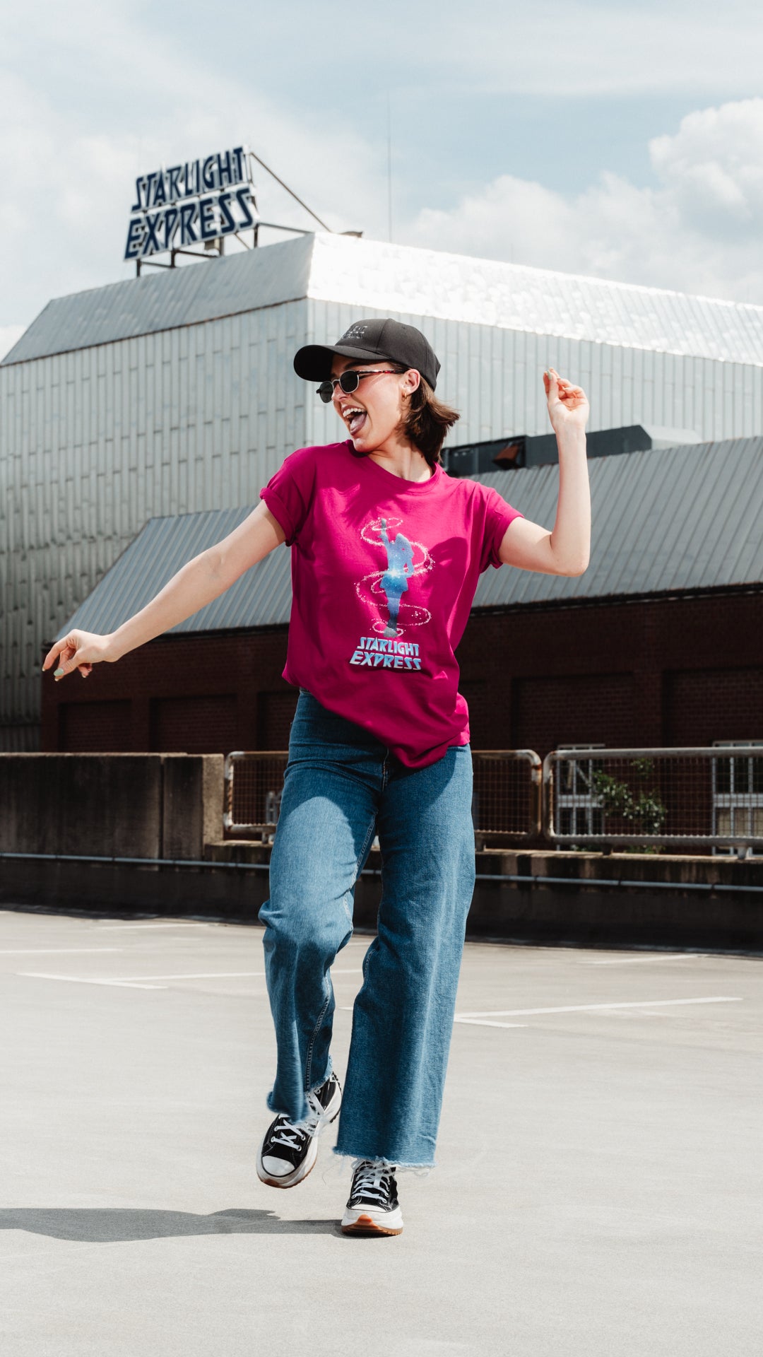Junge Frau tanzt vor dem Starlight Express Theater und trägt ein T-Shirt in Farbe fuchsia mit Pearl Motiv und Starlight Express Logo auf der Vorderseite.